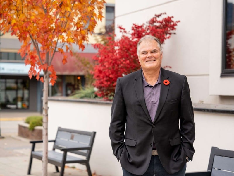 Evan Parliament, an older Caucasian male in a suit standing in front of city hall with a poppy pinned to his suit jacket. He is smiling at the camera with his hands in his pockets looking relaxed.