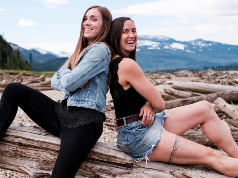 THE STUDIO owners Jacqui Gardiner and Kenley Knock sit back to back on a log, arms crossed staring at the camera with a smile. Both are younger white women with dark brown hair longer than shoulder length,.