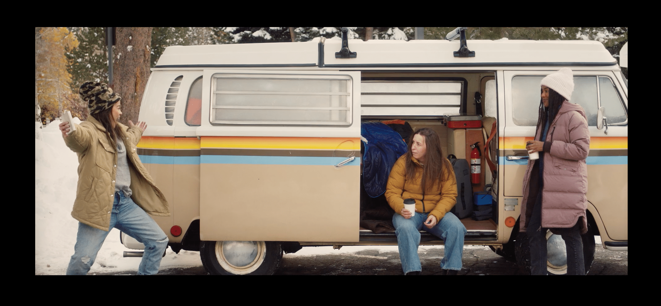 Left to right, Jadyn Wong, Katie Burrell and Chelsea Conwright stand along the side of a camper van in a parking lot filled with snow, all holding a coffee and Jadyn holding a beer in a still from Weak Layers.