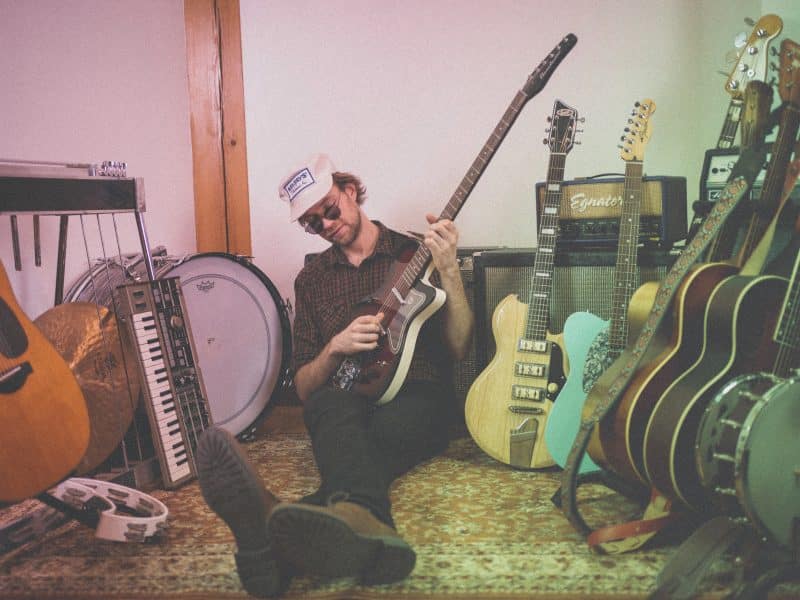 Porch Hounds’ Nik Winnitowy sitting on the floor with his back against an amp. He is playing one electric guitar with a lineup of guitars flanking either side of him.