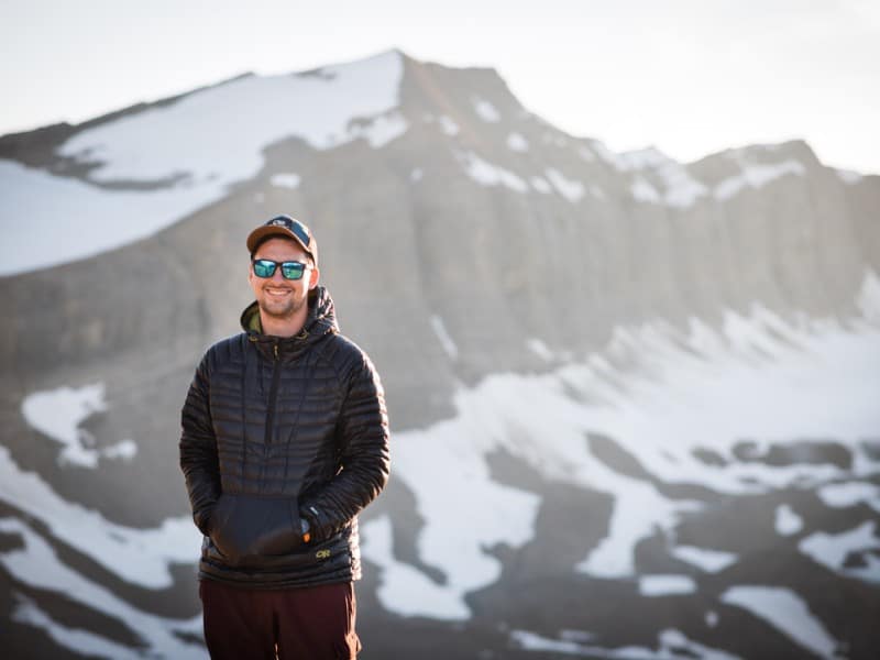Kyle Scott standing in front of a mountain rainge with the sun behind him, wearing sunglasses, a winter jacket and a baseball cap.