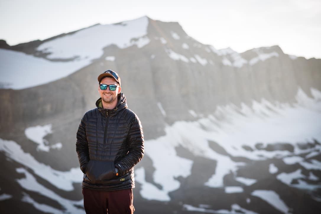 Kyle Scott standing in front of a mountain rainge with the sun behind him, wearing sunglasses, a winter jacket and a baseball cap.