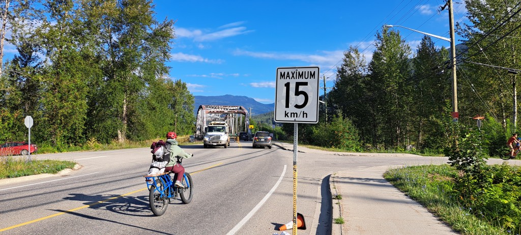 A 15 kmh sign in front of a steel bridge with vehicle and bike traffic on it. The top right steel beam is bent with paint damage also visible.
